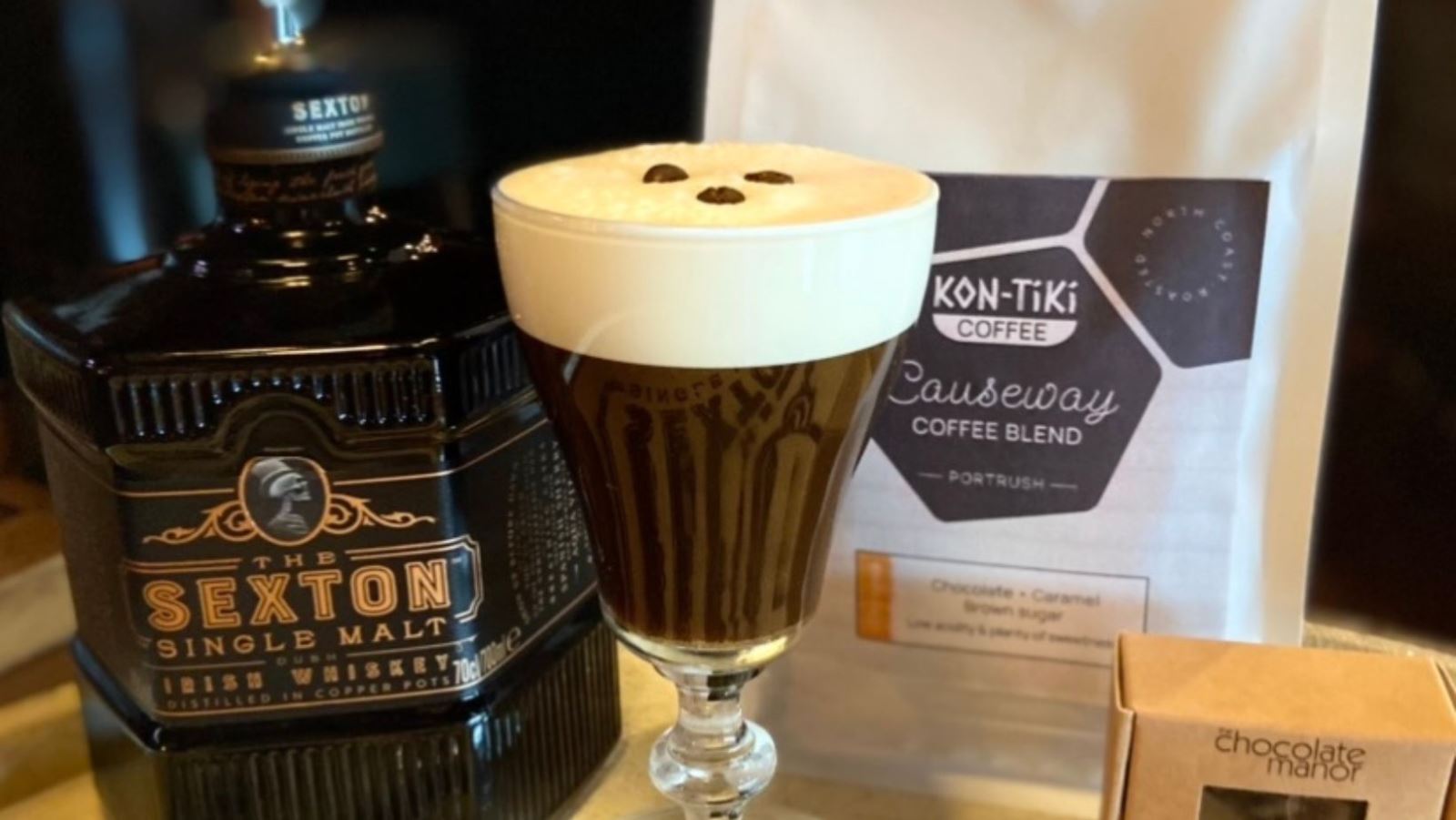 Image of a large Irish coffee in a glass with a bottle of whiskey, chocolates and coffee beside it on the table.